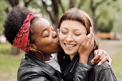 Amorous African female kissing her happy girlfriend on cheek in natural environment