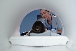 Young female clinician looking at little girl lying on table of mri scan machine before medical procedure