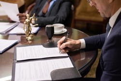Mature businessman or jurist in formalwear signing juridical document by workplace against his colleague looking through papers