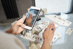 Hand of young woman trying on ring with blue gemstone and taking photograph over display with jewelry