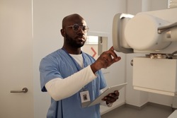 Serious African American male clinician preparing for examination of patient and pressing button on panel of roentgen machine