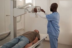 Mature man lying on couch while African American male doctor switching on roentgen machine before examination of patient