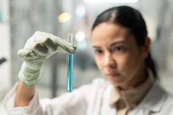 Gloved hand of young female scientist in lab coat holding flask with blue liquid while carrying out scientific experiment in laboratory
