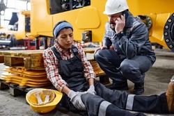 Young female engineer with hurting knee sitting on the floor by anxious male worker in safety helmet and uniform calling ambulance