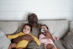 High angle view of exhausted mature African American dad falling asleep while taking care of his baby twin daughters