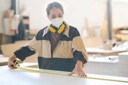Female worker in protective mask using measure tape to make measurements on piece of wood at table