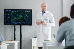 Mature lecturer in white coat with digital tablet standing near the display with human lungs and teaching students at class