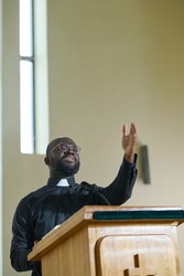 Young priest in black shirt with clerical collar standing by pulpit and speaking in microphone while keeping arm raised in blessing gesture