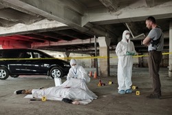Young policeman communicating with forensic expert at crime scene with dead body covered with white sheet in parking area