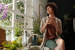 Young serene brunette with cup of coffee looking through window surrounded by plants while sitting by sink in the kitchen