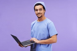 Young successful student or designer in blue t-shirt and beanie hat holding laptop while looking at camera against lavender background