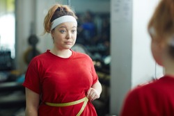 Portrait of cute obese woman standing against mirror in gym and measuring waist size with tape after workout, looking upset and confused