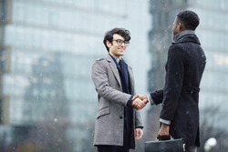 Portrait of two business people, Middle-eastern and African, meeting in snowy city street, smiling, shaking hands and greeting each other