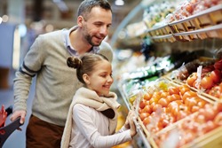 Portrait of cute little girl with dad leaning over vegetable counter choosing fresh ripe tomatoes and other vegetables in supermarket