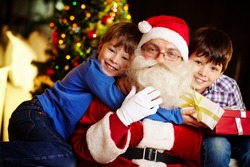 Photo of happy boy embracing Santa Claus with cute kid near by