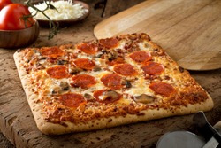 A delicious square crust flatbread pizza with pepperoni, bacon and mushrooms on a rustic wood table top.