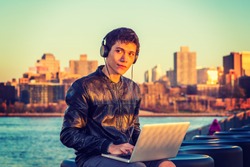 Asian American college student traveling, studying in New York, wearing black leather jacket, headphone, sitting by river in sunset, listening music, working on laptop computer. Instagram effect.
