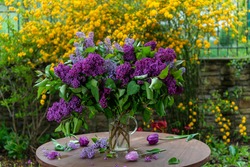 A bouquet of purple lilac in a vase on a white table with yellow flowers in the background