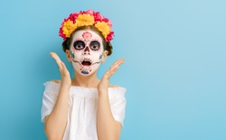 Adorable zombie in flower wreath posing on blue background. Happy child with Halloween creative makeup. Girl celebrating for Mexican Day of the Dead.