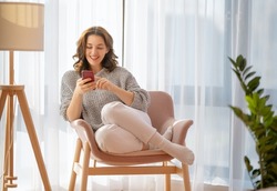 Happy casual beautiful woman is using a phone sitting on armchair at home.