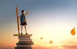 Back to school! Happy cute industrious child standing on the tower of books and holding a huge pencil on background of sunset sky. Concept of education and reading. The development of the imagination.