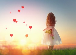 Sweet child girl looking at red balloons. Little child girl holding bouquet of flowers. Balloons in shape of heart flying in the sunset sky. Wedding, Valentine, love concept. 