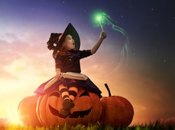 Happy Halloween! Cute cheerful little witch with a magic wand and book of spells. Beautiful child girl in witch costume sitting on the big pumpkin, conjuring and laughing.