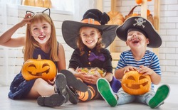 Happy brother and two sisters on Halloween. Funny kids in carnival costumes indoors. Cheerful children play with pumpkins and candy.