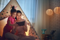 Happy loving family. Young mother and her daughter girl play in children room at the bedtime. Funny mom and lovely child are having fun with tablet.