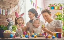 Mother, father and daughters are painting eggs. Happy family are preparing for Easter. Cute little child girl wearing bunny ears.