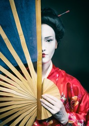 Woman in geisha makeup covering half of her face with a big fan