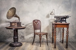 Vintage loft room with old typewriter and lamp on wooden desk, antique chair and classic gramophone with brass horn  front concrete wall background with shadows. Retro style filtered photo