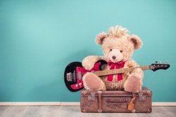 Retro Teddy Bear toy with hairstyle playing bass guitar and sitting on old leather travel luggage. Hard rock or heavy metal music concept. Vintage nostalgia style filtered photo
