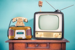 Retro outdated TV set from 60s, old FM radio, golden microphone and classic telephone on oak wooden table front mint blue background. News, journalism nostalgia concept. Vintage style filtered photo