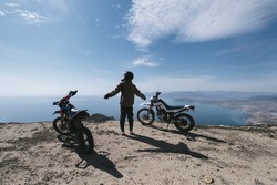 Female motorcyclist relaxing in dirt motorcycle travel on mountain cliff, beautiful sea shore and mountains landscape on background 