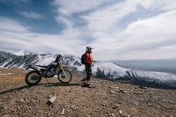 Active man resting near dirt bike looking on beautiful snow mountain landscape