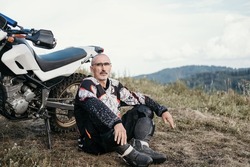 Portrait of 60-year-old Active mature man with grey hair  resting near enduro motorcycle in beautiful mountain landscape