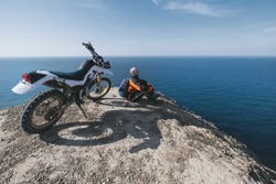 Active elderly man resting near dirt motorcycle on beautiful clff top, sea landscape on background