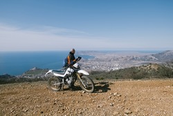 Active elderly man riding dirt motorcycle in beautiful mountains hills with city on sea shore landscape