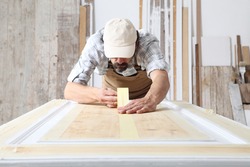 Male carpenter working the wood in carpentry workshop, putting paper masking tape on a wooden door, wearing overall and cap
