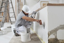 man plasterer construction worker at work, takes plaster from bucket and puts it on trowel to plastering the wall, wears helmet inside the building site of a house