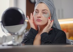 Patches for eyes. Attractive woman in kitchen with towel on her head glues transparent patches to skin under her eyes. Hygiene, beautician, makeup, face care concept.