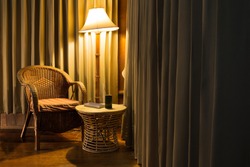Rattan chair and stool under the light from the lamp with bamboo cup and the book surround by beautiful curtain.