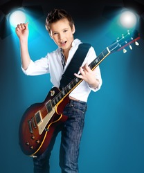 A young boy playing on the electric guitar on the stage with bright blue projector behind him