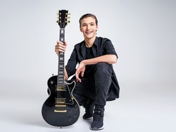 Fifteen years old guitarist with a  black electric guitar. Teenage musician holds guitar, posing at studio. The young caucasian guy with a guitar. Stylish musician. Heavy metal player. 
