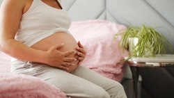 A pregnant girl is sitting on the bed and holding her big belly. A long gestation period.