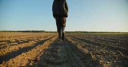 Low angle: man walking in rubber boots in a farmer's field, the blue sky above the horizon. Man walking through an agricultural field. Farmer walks through a plowed field in early spring. 