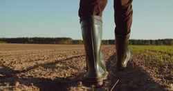 Low angle: man walking in rubber boots in a farmer's field, the blue sky above the horizon. Man walking through an agricultural field. Farmer walks through a plowed field in early spring. 