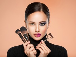 Portrait of a girl with  tools for making makeup near face.  One half face of a beautiful white woman with  bright makeup and the other is natural. Woman holds makeup brush and eyelash curler. Natural