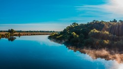 Aerial  view of a beautiful summer  landscape over river while dawn. Top view over river with a smooth water surface reflecting blue sky. Morning evaporation on a river while sunrise. 